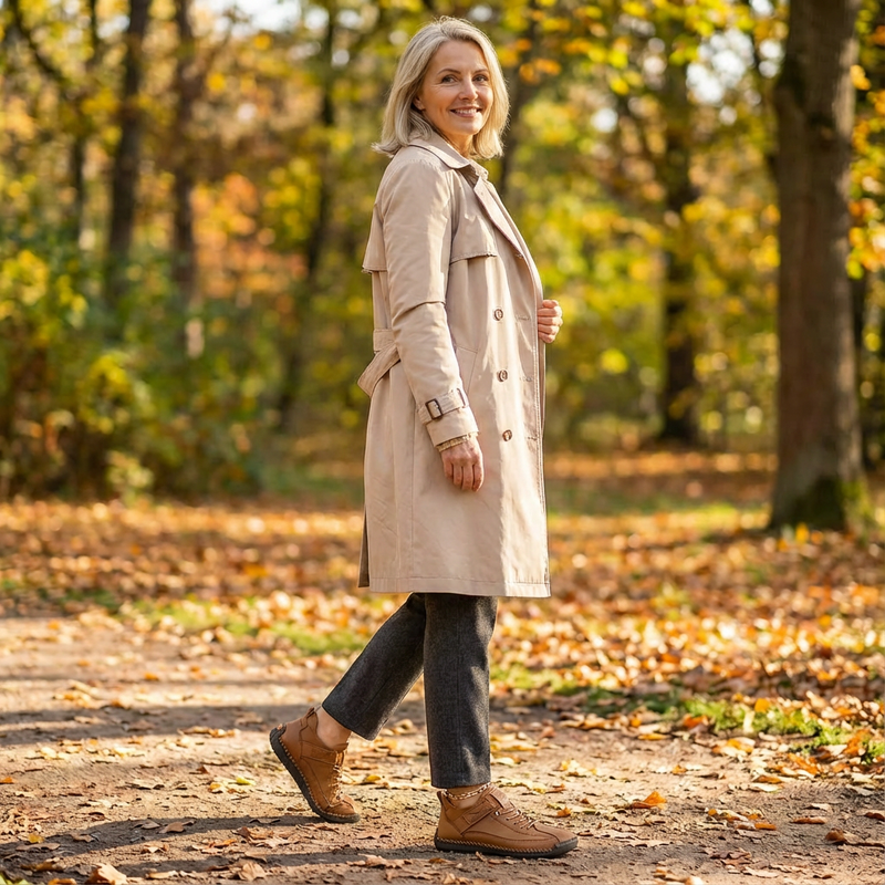 Wandelende vrouw in beige trenchcoat op een bospad vol herfstbladeren, gekleed in bruine leren sneakers met veters. 18