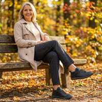 Glimlachende vrouw met beige trenchcoat zit op een houten bankje in herfstkleuren en draagt zwarte sneakers met veters. 17