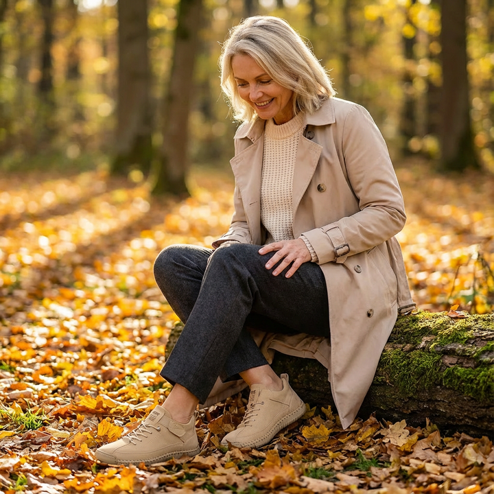 Zittende vrouw in beige trenchcoat op een boomstam in herfstbos, draagt beige sneakers met veters en kijkt glimlachend naar haar schoenen. 15