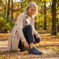 Gehurkte vrouw in beige trenchcoat op een bospad kijkt naar haar donkerblauwe sneakers met veters en klittenbandriempje. 20