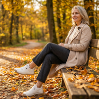 Vrouw met beige trenchcoat zit op een bankje in een herfstpark en draagt witte sneakers met veters, benen over elkaar. 16