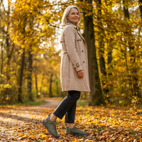 Vrouw in beige trenchcoat loopt over een herfstpad tussen gele bomen en draagt groene sneakers met bruine details. 19