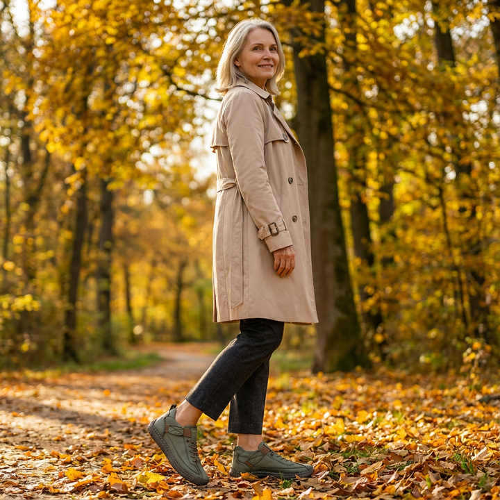 Vrouw in beige trenchcoat loopt over een herfstpad tussen gele bomen en draagt groene sneakers met bruine details. 19
