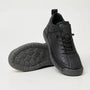 Zwarte sneaker met korrelig oppervlak en donkergrijze zool, met de ronde noppenzool van de tweede schoen in beeld. 3