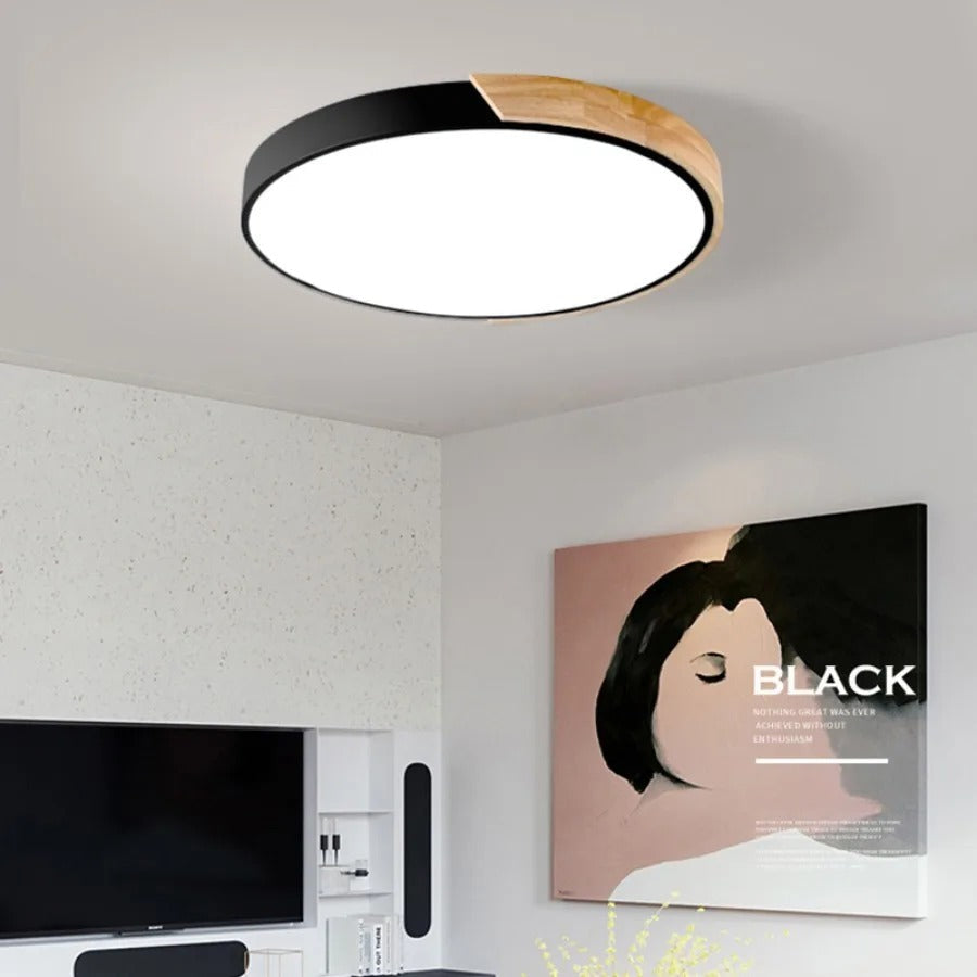 Zwarte ronde plafondlamp met houten accent aan de rand, brandend in een woonkamer boven een tv en een kunstwerk. 1