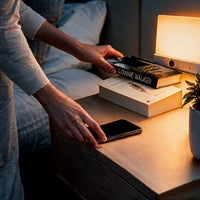 Hand legt een boek op een nachtkastje naast een telefoon, terwijl de lamp warm oplicht 10