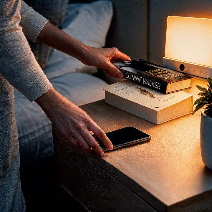 Hand legt een boek op een nachtkastje naast een telefoon, terwijl de lamp warm oplicht 10