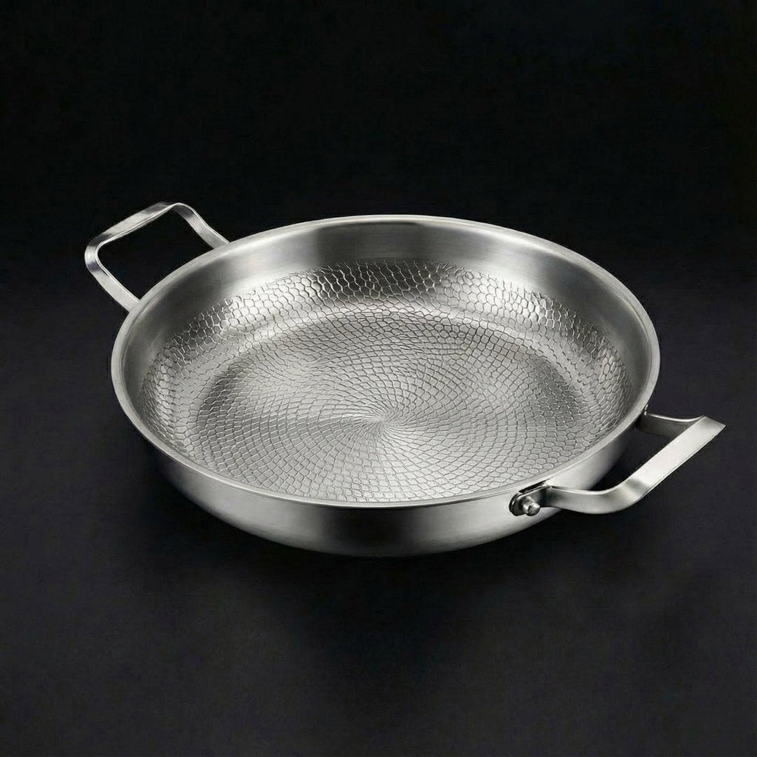 Lege metalen pan met twee handgrepen en een reliëfpatroon op de bodem, schuin van boven op een zwarte achtergrond. 1
