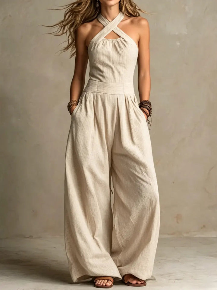 Model draagt de jumpsuit in zandbeige met overslaghalslijn, hoge taille en wijde pijpen, gecombineerd met armbanden en sandalen. 2