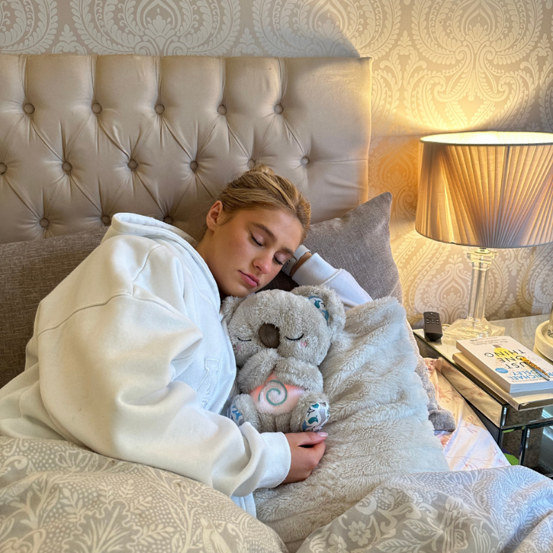 Slapende vrouw in witte hoodie in bed met een grijze koala in haar armen, met een lamp en boeken op het nachtkastje. 14