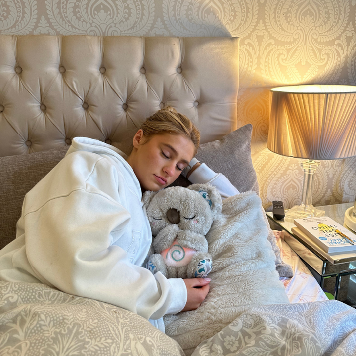 Slapende vrouw in witte hoodie in bed met een grijze koala in haar armen, met een lamp en boeken op het nachtkastje. 14