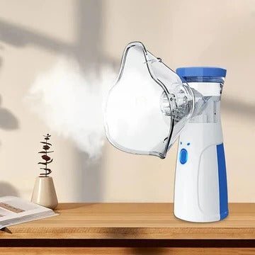 Blauw-witte handvernevelaar met masker op een houten tafel, waaruit een fijne witte nevel komt, naast een boek en een plantje. 1
