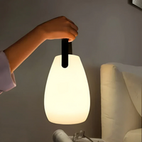 Hand houdt de druppelvormige lamp aan de zwarte lus omhoog naast een bed met bril 1