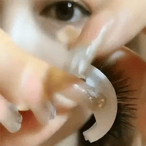 Close-up van een vrouw die met haar vingers een wimperstukje op een wit houdertje vasthoudt vlak bij haar oog. 14