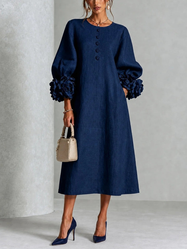 Model draagt de midi-jurk in marineblauw, vooraanzicht, met bloemenknopen voor, ruime mouwen met grote stoffen rozen en een beige handtas. 1