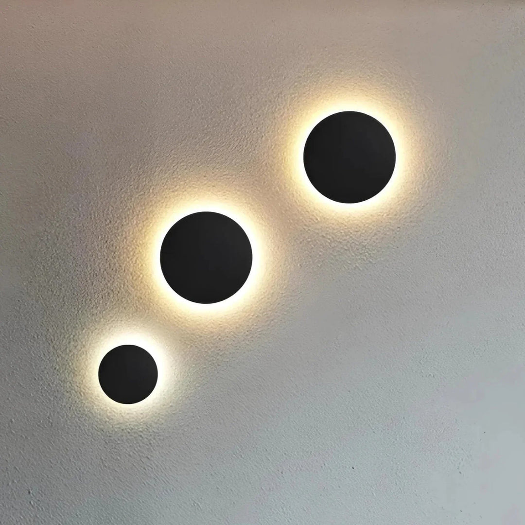 Drie zwarte ronde wandlampen in verschillende maten diagonaal op een lichte muur, met een warme gloed rondom de schijven. 2