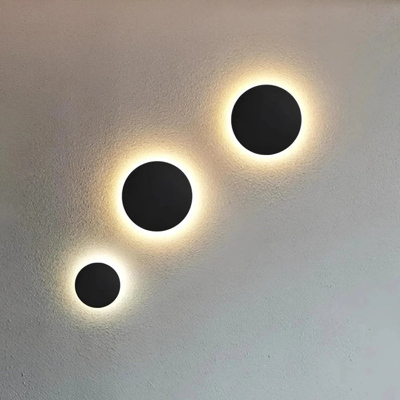 Drie zwarte ronde wandlampen in verschillende maten diagonaal op een lichte muur, met een warme gloed rondom de schijven. 2