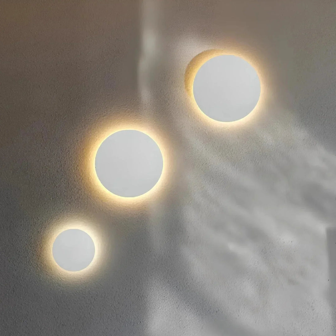 Drie witte ronde wandlampen in verschillende maten diagonaal op een muur, elk met een zachte lichtkrans eromheen. 4