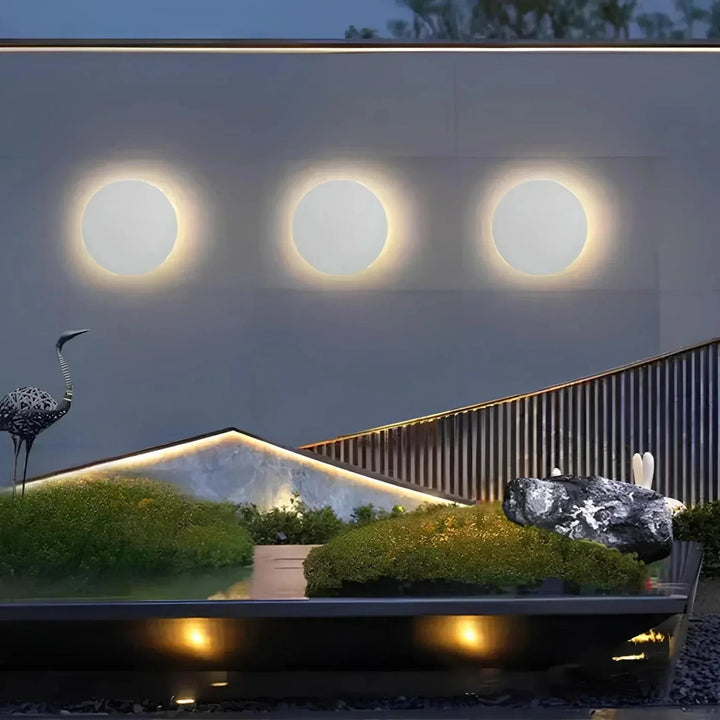 Drie witte ronde wandlampen op een rij aan een buitenmuur boven een moderne tuin met mos, rotsen en een sculptuur. 5