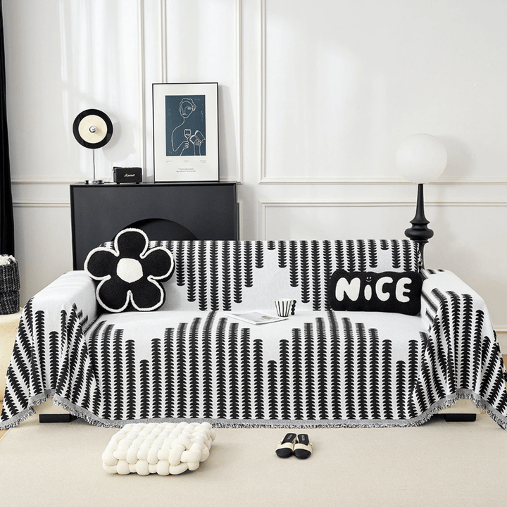 Zwart-wit gestreepte bankhoes met franjes over een bank, gestyled met bloemvormig kussen, tekstkussen, tijdschrift en kopje in een witte kamer. 1