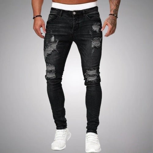Voorkant van slim fit jeans met scheuren in zwart verweerd denim, gedragen door een model met witte sneakers. 1