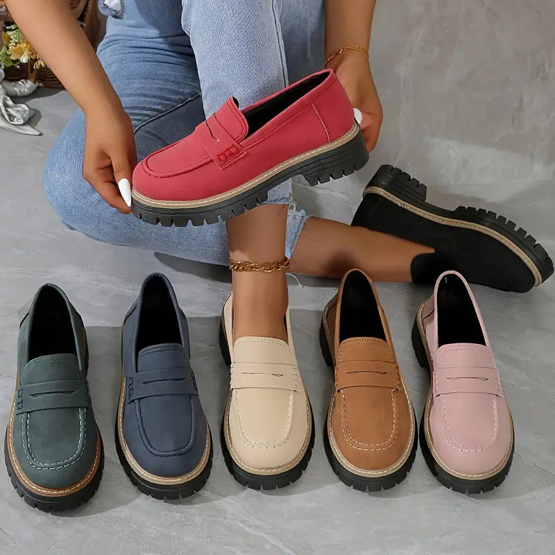 Loafers in zes kleuren op een grijze vloer, waarbij een model een rode loafer omhoog houdt. 2