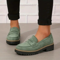 Groene suèdelook loafers met dikke zwarte profielzool, gedragen onder zwarte opgerolde broekspijpen voor een witte bakstenen muur. 1