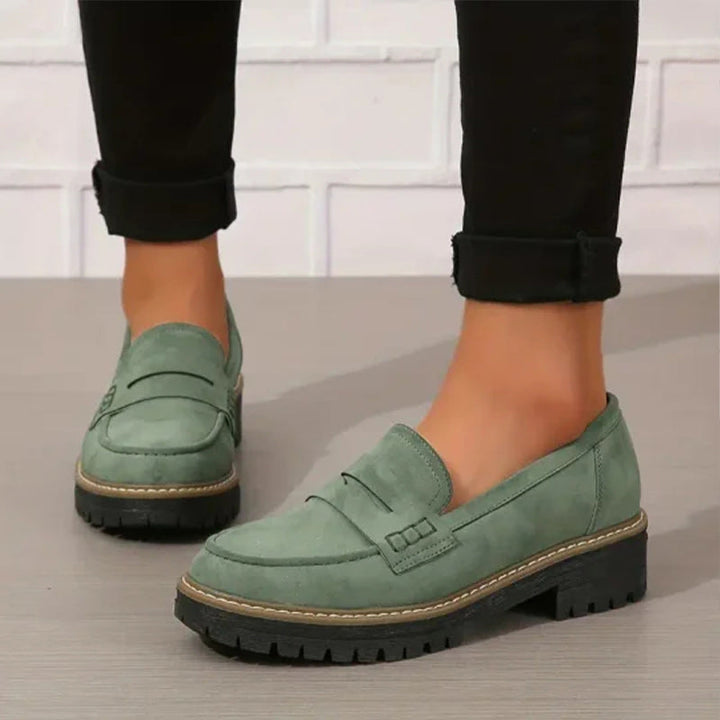 Groene suèdelook loafers met dikke zwarte profielzool, gedragen onder zwarte opgerolde broekspijpen voor een witte bakstenen muur. 1