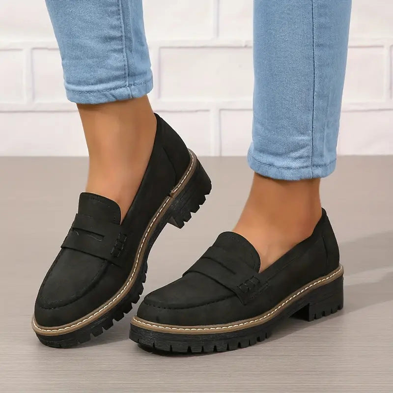 Zwarte loafers met dikke profielzool gedragen onder opgerolde blauwe jeans, één voet schuin gedraaid. 6