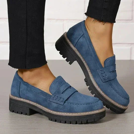Blauwe suèdelook loafers met zwarte profielzool gedragen met zwarte broek, één voet opgetild met zijaanzicht van de zool. 9