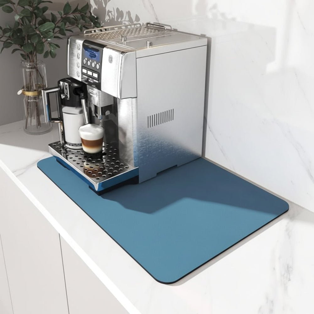Blauwe afgeronde mat onder een zilverkleurig koffieapparaat op een wit marmeren aanrechtblad, met een cappuccino in het glas. 1