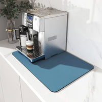 Blauwe afgeronde mat onder een zilverkleurig koffieapparaat op een wit marmeren aanrechtblad, met een cappuccino in het glas. 1