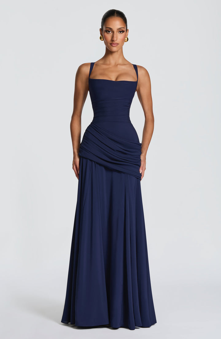 Vooraanzicht van de marineblauwe maxi-jurk op een model, met vierkante halslijn, geplooide taille, heupdrapering en ruim uitlopende rok. 7