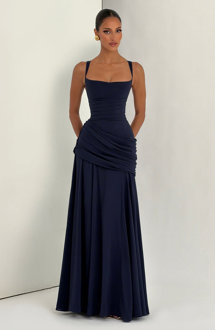 Model in de marineblauwe maxi-jurk voor een lichte muur, vooraanzicht met gedrapeerde heupen en een lange, ruim vallende rok. 13