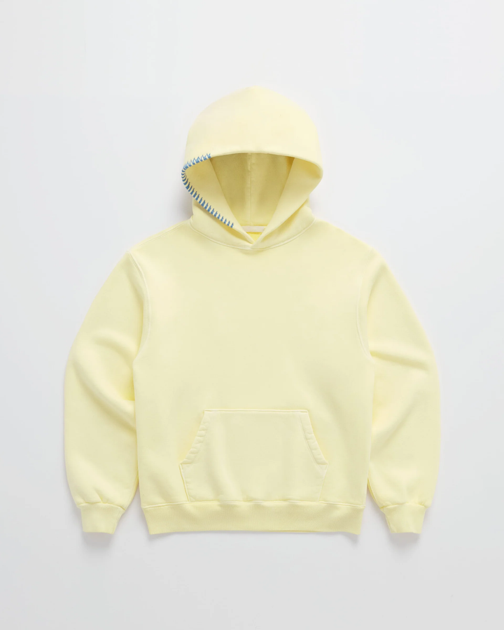 Hoodie in zachtgeel van voren op een lichte achtergrond, met blauwe stiksels langs de rand van de capuchon en een buidelzak. 1
