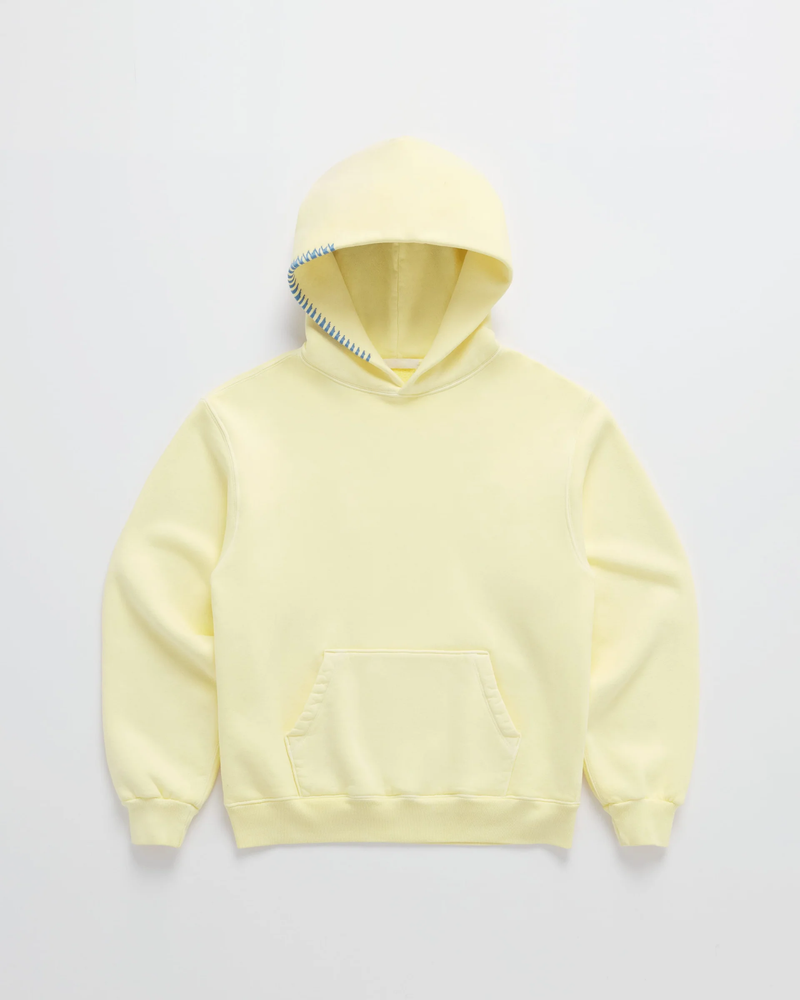 Hoodie in zachtgeel van voren op een lichte achtergrond, met blauwe stiksels langs de rand van de capuchon en een buidelzak. 1