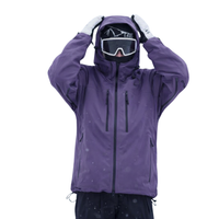 Paarse snowjack gedragen door een model met skibril en beide handen op de capuchon, vooraanzicht met borstzakken met ritsen. 1