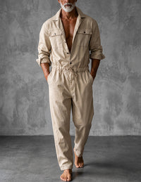 Man in beige overall met opgerolde mouwen, open kraag en trekkoord, blootsvoets voor een grijze betonnen muur. 1