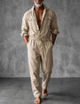 Man in beige overall met opgerolde mouwen, open kraag en trekkoord, blootsvoets voor een grijze betonnen muur. 1