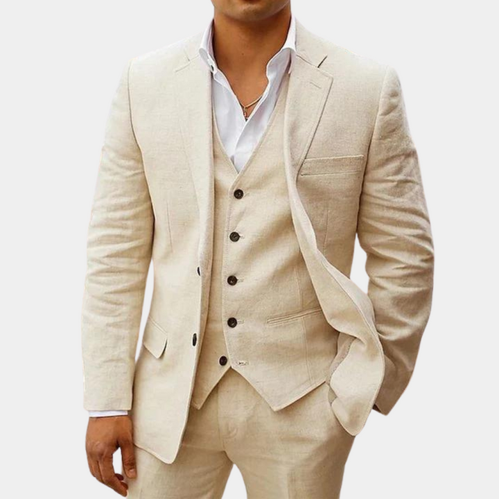 Model draagt het pak in beige met wit overhemd, gilet met vijf knopen en een hand in de broekzak. 1