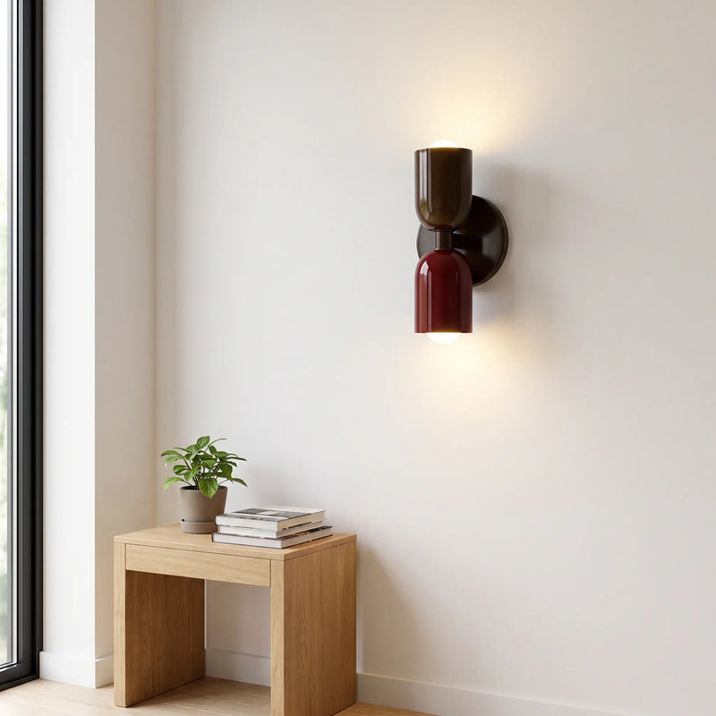 Wandlamp in donkerbruin en bordeauxrood, brandend aan een witte muur boven een houten krukje met plant en boeken. 10