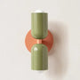 Wandlamp met twee glanzende groene kappen op een ronde oranje wandplaat, tegen een lichte muur. 21