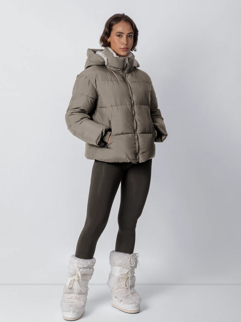 Model in taupegroen pufferjack met dichte rits, donkere legging en witte pluizige snowboots, handen in de zakken 21