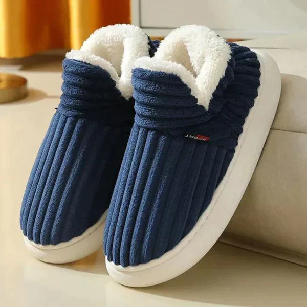 Marineblauwe geribbelde pluchen pantoffels met witte fleece voering en dikke witte zool, schuin neergezet naast een bank. 1