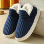 Marineblauwe geribbelde pluchen pantoffels met witte fleece voering en dikke witte zool, schuin neergezet naast een bank. 1
