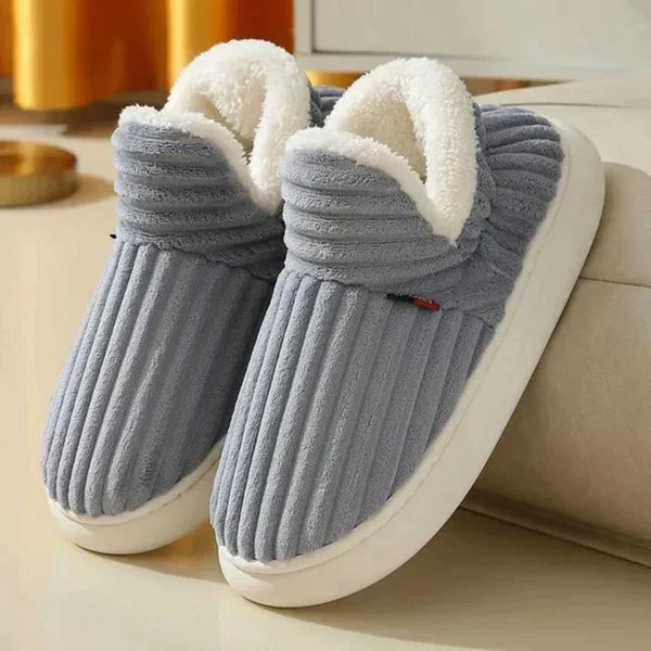 Grijze geribbelde pluchen pantoffels met witte fleece voering en witte zool, schuin neergezet naast een bank. 2