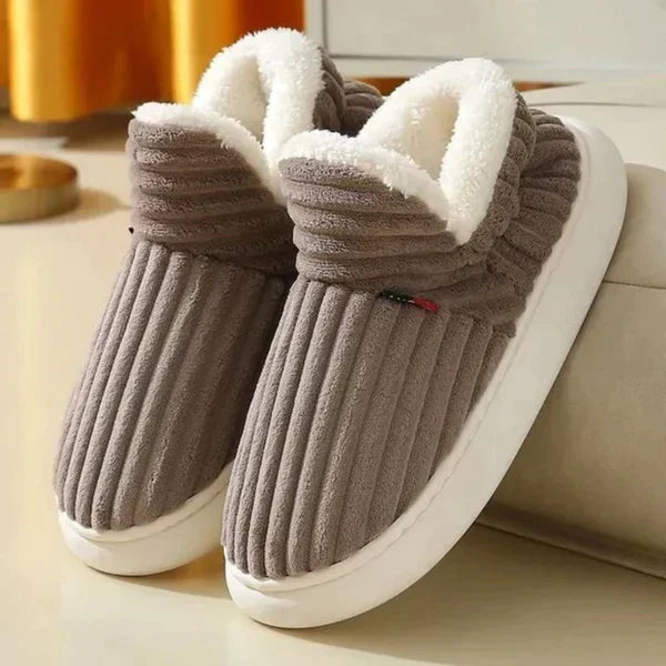 Taupebruine geribbelde pluchen pantoffels met zachte witte fleece rand en witte zool, schuin neergezet naast een bank. 3