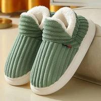 Groene geribbelde pluchen pantoffels met witte fleece voering en witte zool, schuin neergezet naast een bank. 4