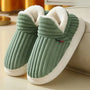 Groene geribbelde pluchen pantoffels met witte fleece voering en witte zool, schuin neergezet naast een bank. 4