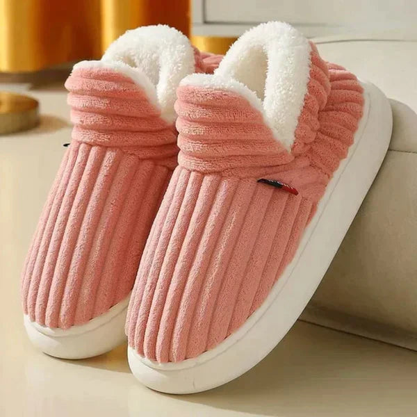 Roze geribbelde pluchen pantoffels met witte fleece voering en dikke witte zool, schuin neergezet naast een bank. 5